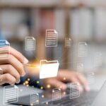 A person is typing on a laptop with a bunch of papers coming out of it.